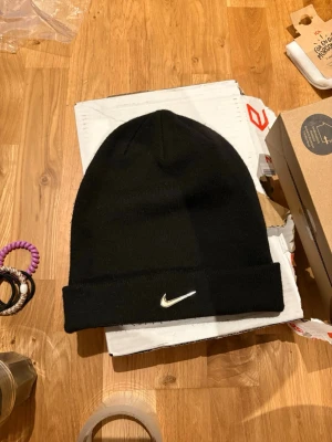 Svart mössa från Nike med metall-logga - Svart mössa från Nike med en snygg metallfärgad swoosh-logga framtill. Mössan är ribbstickad och har en uppvikt kant för extra stilpoäng. Perfekt för dig som vill ha en clean och sportig look under kyliga dagar.