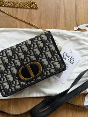 Dior axelväska med monogram och kedja - Säljer en snygg axelväska från Dior med klassiskt monogrammönster i svart och beige. Väskan har guldfärgad kedja, magnetlås och detaljer i svart skinn. Framsidan pryds av ett stort CD-emblem i guld och svart. Perfekt storlek för det viktigaste och har flera fack inuti.