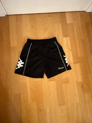 Svarta träningsshorts från Kappa - Svarta sportiga shorts från Kappa med vita loggor på sidorna och kontrastsömmar. Shortsen har elastisk midja med snörning. Perfekta för träning eller sportaktiviteter.
