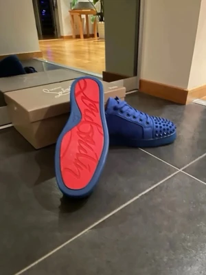 Louboutin  - Säljer ett par blå sneakers från Christian Louboutin med coola nitar på tån och röd sula med logga. Skorna har snörning och är gjorda i mocka med beige insida. Perfekt för dig som vill sticka ut med en lyxig och unik stil.