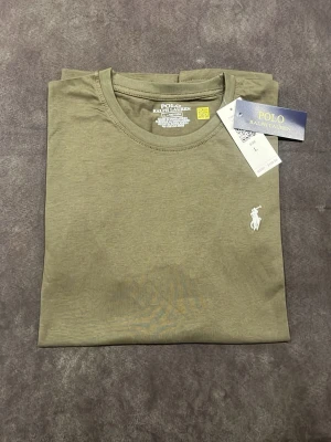 Olivgrön t-shirt från Polo Ralph Lauren - Snygg olivgrön t-shirt från Polo Ralph Lauren med klassisk rund hals och korta ärmar. T-shirten har en vit broderad logga på bröstet och är gjord i mjuk bomull som känns skön mot huden. Perfekt för dig som gillar stilrena och enkla plagg med premiumkänsla.