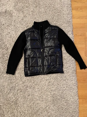 Svart pufferjacka All Caps One - Svart pufferjacka från All Caps One med glansig, quiltad framsida och stickade ärmar. Jackan har dragkedja framtill och två sidofickor med dragkedja. Snygg hög krage och broderad logga på ärmen. Perfekt för dig som gillar streetstyle.