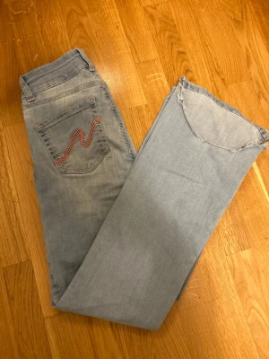 Ljusblå jeans från Nelly med broderad ficka - Säljer ett par ljusblå jeans från Nelly med raka ben och råa, fransiga benslut. Jeansen har en unik bakficka med rödvitt broderat mönster och rosa sömmar som ger en cool detalj. Perfekta för dig som gillar en avslappnad och trendig look. jag har försökt förkorta de men gav upp , det går att fixa