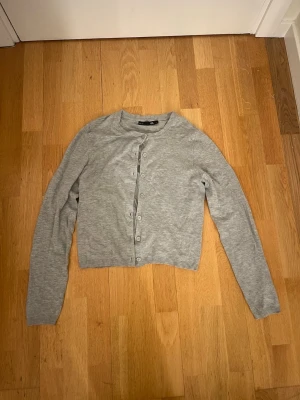 Grå kofta från 157 med knappar - Snygg grå kofta från 157 i storlek L. Modellen är klassisk med rund hals, långa ärmar. Koftan har knäppning framtill med matchande grå knappar. Perfekt att slänga på sig när det är lite kyligt ute.