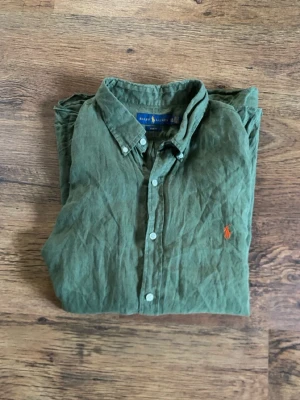 Grön linneskjorta från Ralph Lauren - Snygg grön skjorta från Ralph Lauren i slim fit-modell, tillverkad i linne. Klassisk krage, vita knappar och den ikoniska orangea broderade loggan på bröstet. Perfekt för dig som gillar stilrena och bekväma plagg med lite extra attityd. XXL men sitter tajt.