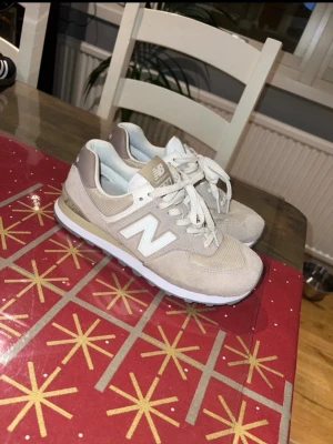 Beige New Balance 574 sneakers - Säljer ett par snygga New Balance 574 sneakers i beige och ljusgrå toner. Skorna har klassisk snörning, rund tå och platt sula. Ovandelen är i mocka och mesh med vita detaljer och logga på sidan. Perfekta för dig som gillar stilrena och bekväma sneakers.