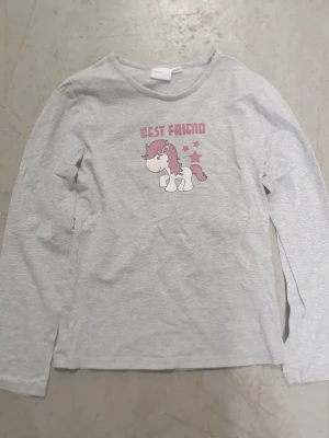 Tröja - Säljer en grå långärmad tröja från mywear young i storlek 134/140. Tröjan har ett tryck med en söt unicorn och texten 'Best Friend' i rosa samt rosa stjärnor. Materialet är mjuk bomull och passformen är normal. Perfekt för dig som gillar gulliga motiv!