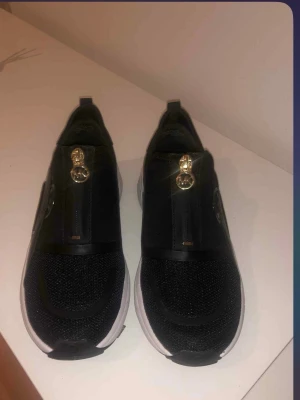 Svarta Michael Kors sneakers med gulddetalj - Snygga svarta sneakers från Michael Kors med vit och svart platt sula. Ovandelen är i mesh och syntet, och har en guldfärgad MK-detalj på ovansidan. Skorna har en modern, sportig look och är slip-on utan snörning. Perfekta för dig som gillar stilrena och trendiga sneakers.