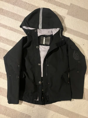 Vindjacka från Sail Racing - Svart vindjacka från Sail Racing med huva och grått meshfoder. Jackan har dragkedja, knappar och logodetaljer på ärm och bröst. Praktiska fickor med lock och justerbar nederkant. Storlek S. 