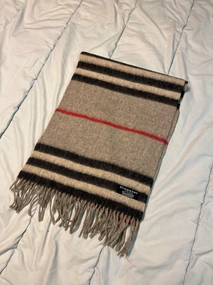 Randig halsduk från Burberry i kashmir - Säljer en klassisk halsduk från Burberry i 100% kashmir. Den är grå med svarta, vita och röda ränder samt fransar i ändarna. Perfekt accessoar för att lyfta din outfit med en tidlös och lyxig känsla. N15
