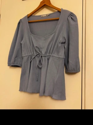 Ljusblå blus med knut från Lager157 - En väldigt fin ljusblå blus från Lager157 i storlek S. Blusen har trekvartsärm med ett knut vid midjan för en perfekt fin och feminin form. Passar perfekt till våren och sommaren.