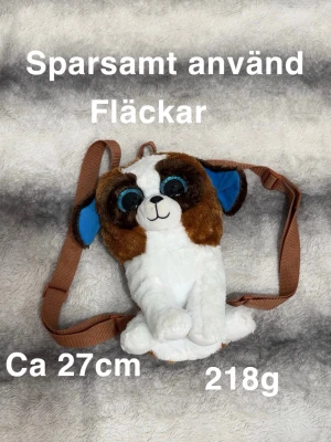 Ryggsäck med hundmotiv - Supergullig ryggsäck formad som en hund med stora blå ögon. Axelremmarna är bruna och justerbara. Perfekt storlek för att bära småsaker, ca 27 cm lång och väger 218g. Söt accessoar för dig som gillar djur och unika väskor.🌸Fläckar. Sparsamt använd.