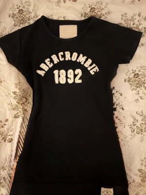 Mörk blå t-shirt Abercrombie 1892 - Mörk blå t-shirt från Abercrombie & Fitch med vit text 'Abercrombie 1892' på bröstet och en snygg patch längst ner. Klassisk rund hals och korta ärmar, tillverkad i mjuk bomull som är skön mot huden. Perfekt för dig som gillar stilrena och 2000s plagg.Storleken är S-M