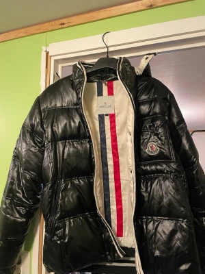 (Behöver kemtvätt) - Svart pufferjacka med glansig finish och ikonisk logga på bröstet. Insidan har vita paneler med röda och marinblå ränder samt en unik serietidningsetikett. Jackan har dragkedja, knappar och ribbade muddar. Perfekt för dig som vill ha en snygg och varm jacka. 