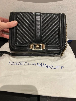 Rebecca Minkoff väska - Snygg svart cross body bag från Rebecca Minkoff i skinn med quiltad framsida och guldfärgad kedja. Väskan har ett elegant mönster med diagonala ränder och ett stilrent metallspänne framtill. Perfekt storlek för telefon, plånbok och nycklar. Guldiga färgen på metalldelarna är något sliten. Skinnet är även det slitet i kanterna. Helhetsintrycket är ändå bra.
