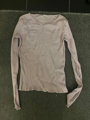 Ljuslila ribbad långärmad topp - Säljer en ljuslila långärmad topp med ribbad struktur och v-ringad hals. Toppen har en slim passform och är tillverkad i ett mjukt, stretchigt material som sitter skönt mot kroppen. Perfekt att styla med jeans eller kjol för en enkel och snygg look.