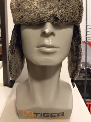 Grå Pälsmössa från Handskkompaniet - Storlek M  - Säljer en snygg grå pälsmössa från Handskkompaniet. Mössan har mjuk konstpäls på utsidan och quiltad svart insida med spänne under hakan. Perfekt för kalla dagar när du vill hålla stilen.