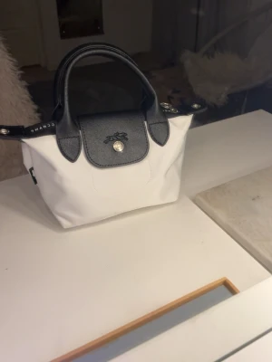 Vit och svart handväska från Longchamp - Säljer en stilren handväska från Longchamp i vit textil med svarta detaljer och handtag i skinn. Väskan har ett klassiskt, kompakt format med silverfärgad knapp framtill och präglad logga på locket. Perfekt för dig som gillar minimalistisk design och snygga kontraster.