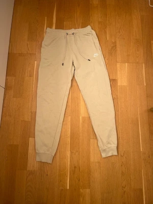 Beige mjukisbyxor från Nike - Säljer ett par beige mjukisbyxor från Nike med dragsko i midjan och klassisk logga på ena benet. Byxorna har en avslappnad passform och är perfekta för chill dagar eller när du vill vara bekväm.