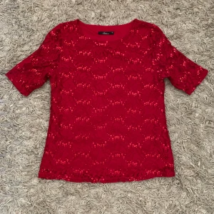 Röd paljettblus med spetsdetaljer - Supersnygg röd blus med kort ärm, täckt av glittrande paljetter och blommig spets. Perfekt för dig som vill sticka ut med en festlig vibe. Blusen har rund halsringning och är lätt transparent i ärmarna tack vare spetsen.