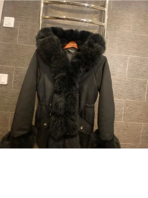 Svart pälsjacka från Hollies - Säljer en svart pälsjacka från Hollies med fluffig fuskpäls längs krage, ärmslut och framkant. Jackan har snygg och markerad siluett med knappar och fickor framtill. 