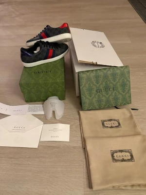 Gucci sneakers med röd och blå detalj - Säljer ett par svarta Gucci sneakers med klassiskt GG-mönster, vita platta sulor och snörning. Skorna har röd och blå rand på sidan samt metallicröd och metallicblå häl med Gucci-logga. Materialet är skinn och textil. Kommer med originalkartong och dustbags.