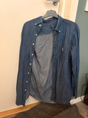 Blå skjorta från GANT i bomull - Säljer en klassisk blå skjorta från GANT i storlek S. Skjortan har lång ärm, bröstficka med liten logga och vita knappar hela vägen. Tillverkad i mjuk bomull och har en avslappnad passform som funkar till många olika stilar.