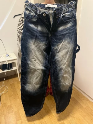 Blå baggy jeans med blekta detaljer - Säljer ett par blå baggy jeans med tydliga blekta partier framtill och baktill. Jeansen har breda ben, stora fickor och hällor för bälte. Materialet är klassisk jeans och passformen är loose för en avslappnad stil. Lite slitna vid fötterna