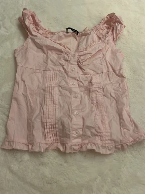 Rosa blus med volang från Brandy Melville - Söt ljusrosa blus från Brandy Melville med volangdetaljer vid axlar och nederkant. Blusen har knappar framtill, rynkade axelpartier och dekorativa veck. Tillverkad i mjuk bomull, perfekt för vår och sommar. 🥰