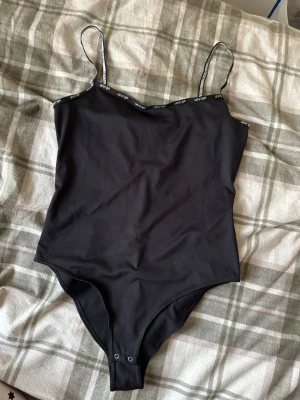 Svart bodysuit från Levi's med smala band - Säljer en svart bodysuit från Levi's med tunna axelband och tryckknappar i grenen. Toppen har logga längs kanten och är i ett mjukt, stretchigt material som sitter tight mot kroppen. Perfekt att styla med jeans eller kjol.