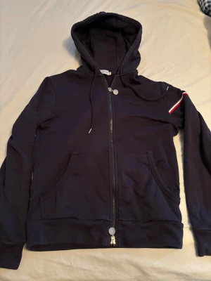 Mörkblå Moncler hoodie med dragkedja - Snygg mörkblå hoodie från Moncler med hel dragkedja, huva och snörning. Tröjan har Moncler-logga på ärmen och en röd, vit och blå randdetalj. Tillverkad i mjuk bomull och har kängurufickor framtill. Perfekt för dig som gillar stilrena streetwear-plagg. Köpt från merchsweden