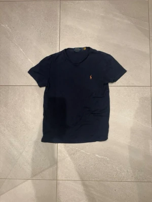 Mörkblå t-shirt från Polo Ralph Lauren - Säljer en mörkblå t-shirt från Polo Ralph Lauren med klassisk v-ringning och korta ärmar. T-shirten har den ikoniska broderade loggan i orange på bröstet. Skön och mjuk bomull som passar perfekt till jeans eller shorts.