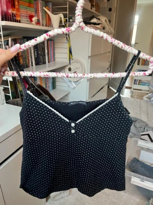 Svart prickig linnetopp från Brandy Melville - Supersöt svart linnetopp med vita prickar från Brandy Melville. Toppen har smala axelband, v-ringning med spetskant och två dekorativa knappar framtill. Perfekt för varma dagar och enkel att styla med jeans eller kjol.