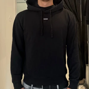 Svart hoodie från Hugo med huva - Svart hoodie från Hugo med minimalistisk vit logga på bröstet. Klassisk modell med huva och snörning, ribbade muddar och mjukt material