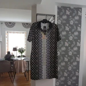 Svart kortklänning  - Svart kortklänning från L√ med vit monogramprint och bred dekorativ rand framtill och baktill. Klänningen har korta ärmar, krage och tydlig L√-logga i guld och beige. Snygg och trendig design som sticker ut.