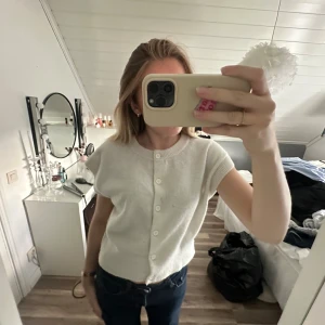 Vit kortärmad kofta med knappar - Supersöt vit kofta med korta ärmar och rund hals. Den har knäppning framtill med vita knappar och är stickad i ett mjukt material. Perfekt att slänga över en topp eller bära som den är för en clean look. Tyvärr har jag råkat spilla något som ej går bort🤩men syns inte tydligt om man har den öppen 