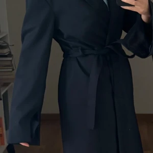 Mörkblå kappa från Filippa K med bälte - Snygg blå kappa från Filippa K med klassisk krage och knytbälte i midjan. Modellen är lång med raka ärmar och har en stilren, minimalistisk look. Perfekt för dig som gillar enkel och tidlös design. Materialet är mjukt och har en lätt glansig finish. 