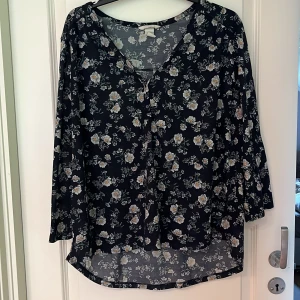 Blus från HM - Svart blus från H&M i storlek XL. Tunn och lätt i materialet med avslappnad passform och rund halsringning. Blusen har 3/4-ärmar och en något längre bakdel vilket gör den bekväm och snygg till både jeans och tights.  Mönstret består av små blommor i ljusa toner (vit, beige och grå) som ger en romantisk och feminin känsla mot den svarta bakgrunden.