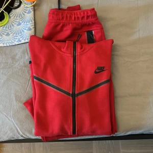 Röd Nike tech fleece  - Säljer ett rött träningsset från Nike med matchande zip-tröja och byxor. Tröjan har svarta detaljer och dragkedja framtill, samt Nike-logga på bröstet. Byxorna har snörning i midjan och Nike-logga på benet. Perfekt för gymmet eller chill hemma. Säljer koftan och byxorna för endast 750kr. Använd par gånger