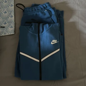 Blåa mjukisbyxor från Nike - Snygga blå mjukisbyxor från Nike med vit logga på benet och dragsko i midjan. Byxorna har en sportig look och är perfekta för chill eller träning. Tillverkade i mjukt material för maximal komfort. Säljer koftan med byxorna för endast 700kr