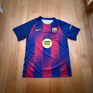 FC Barcelona Nike fotbollströja M - Säljer en officiell FC Barcelona matchtröja från Nike i storlek M. Tröjan har klassiska blå och vinröda vertikala ränder, klubbmärke på bröstet, Nike-logga och sponsorer i gult. Tillverkad i Dri-FIT material som andas och är perfekt för fotbollsträning.