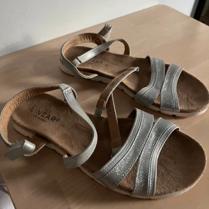 Silvriga sandaler från Linear - Snygga sandaler från Linear med flera silvriga remmar och justerbar kardborre runt ankeln. Använda fåtal gånger, inga defekter.