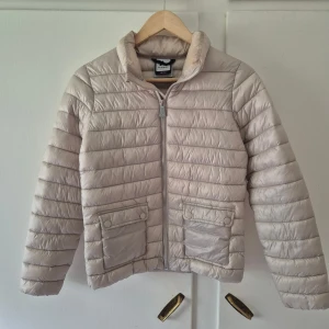 Beige/vit lättviktsjacka från McKinley - Snygg beige/vit lättviktsjacka från McKinley med horisontella sömmar, två fickor framtill med lock och tryckknappar samt dragkedja hela vägen upp. Jackan har en klassisk krage och är perfekt för kyligare dagar.