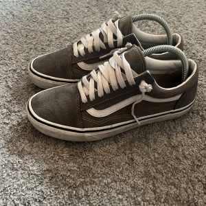 Gråa Vans Old Skool sneakers - Säljer ett par klassiska Vans Old Skool sneakers i grå mocka och skinn med vit sula och vit sidostrip. Skorna har vit snörning och låg profil, perfekt för en avslappnad streetstil. Ikonisk design med rund tå och platt sula.