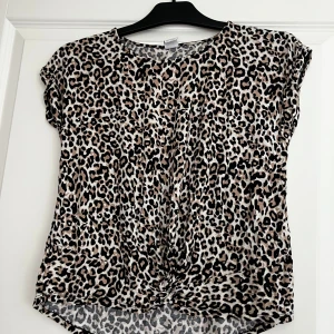 Leopardmönstrad topp från Lindex Kids - Cool topp från Lindex Kids med leopardmönster i beige, svart och brunt. Modellen har rund hals, korta ärmar och en twistad detalj framtill. Perfekt för dig som vill sticka ut med ett djurmönster och en avslappnad passform.