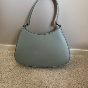 Ljusblå axelväska i skinnimitation - Snygg ljusblå axelväska med minimalistisk design och mjukt rundad form. Väskan har ett brett axelband och silvriga metalldetaljer. Aldrig använd.