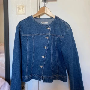 Jeans jacka från gina tricot - Snygg jeansjacka i klassisk blå denim från från gina . Jackan har en unik asymmetrisk knäppning med silvriga knappar och rundad halsringning. Långärmad modell med raka linjer och cool, modern känsla. Perfekt för dig som vill sticka ut lite extra.