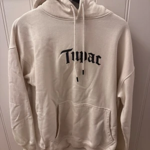 Beige/vit Tupac hoodie med tryck - Säljer en beige/vit hoodie med stor svart Tupac-tryck både fram och bak. Tröjan har huva med snörning och en stor magficka. Oversized passform och mjukt material.