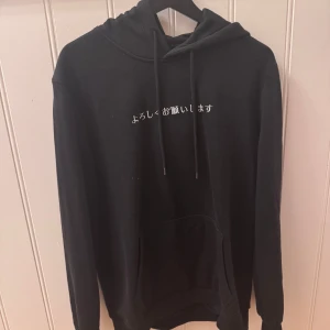 Svart hoodie med japanskt tryck - Svart hoodie med japansk text framtill och en stor vit grafisk illustration av koi-fiskar på ryggen. Tröjan har huva med snörning och en klassisk känguruficka. Perfekt för dig som gillar streetstyle och unika detaljer.