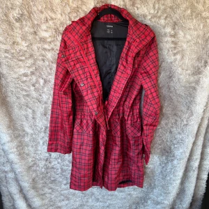 Rutig röd regnjacka från Esmara - Snygg röd regnjacka från Esmara med svart rutmönster, huva och dragsko i midjan. Jackan har både dragkedja och knappar framtill samt stora fickor. Perfekt för regniga dagar och har en cool streetstil.